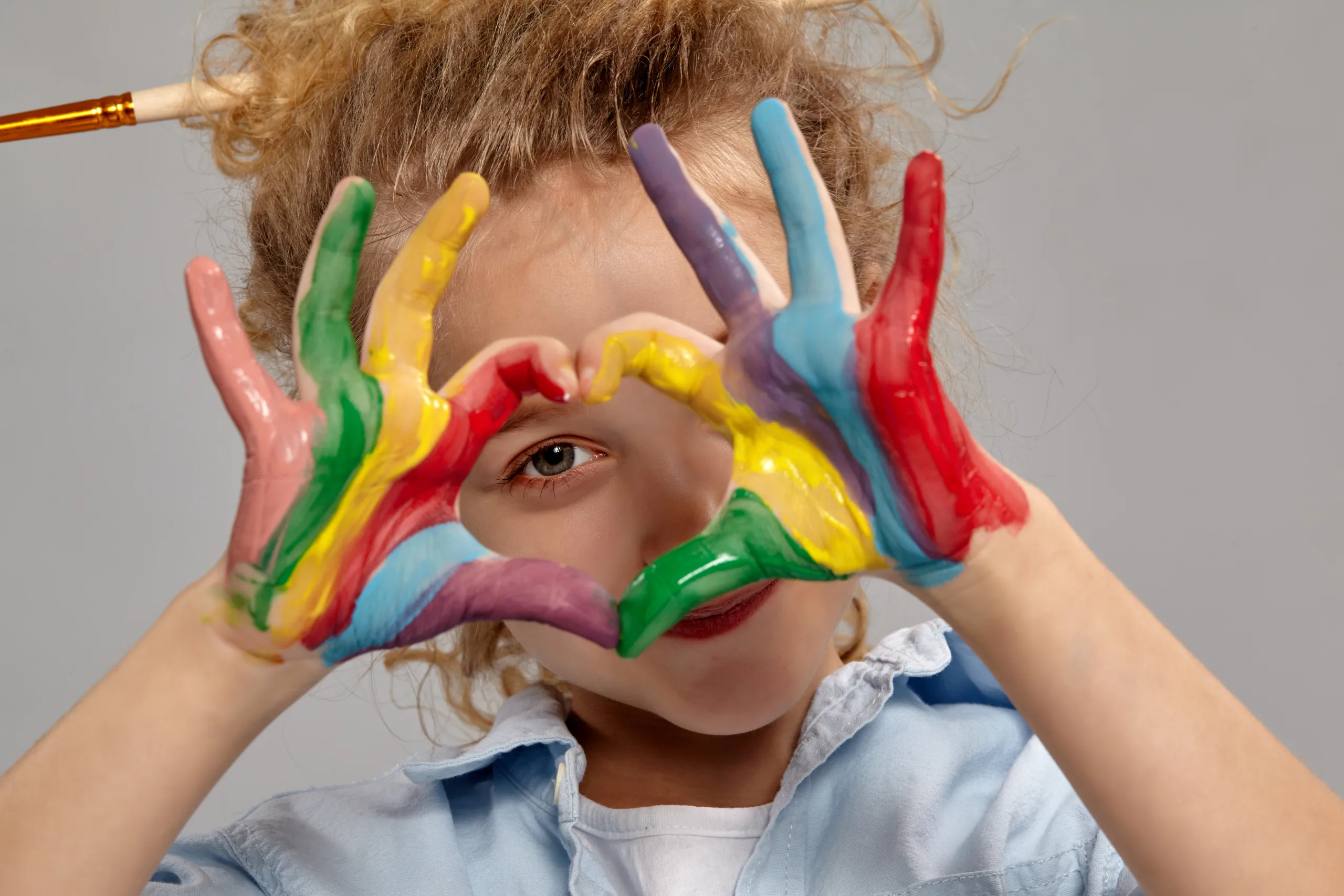 close up shot funny little girl having brush her chic curly blond hair wearing blue shirt white t shirt she is looking through painted hands folded shape heart s scaled